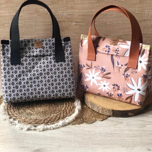 K&ouml;nnte beinhalten: Zwei Einkaufstaschen mit unterschiedlichen Mustern. Eine Tasche ist schwarz mit einem wei&szlig;en geometrischen Muster und die andere ist rosa mit einem wei&szlig;en Blumenmuster. Beide Taschen haben braune Ledergriffe.