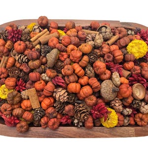 May include: A wooden oval bowl filled with a decorative mix of small orange pumpkins, brown pinecones, cinnamon sticks, large acorns, and bright yellow and deep red dried floral elements, creating a dense, textured autumn-themed arrangement.