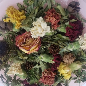 May include: A collection of dried flowers and foliage, including a sunflower, rose, and carnations, in shades of yellow, red, and cream, with green leaves and small pine cones. The arrangement is presented on a pink surface.