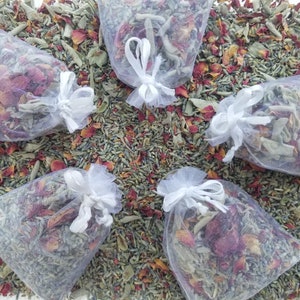 Puede incluir: Cinco pequeñas bolsas de malla blanca llenas de lavanda seca, pétalos de rosa y otras hierbas. Las bolsas están atadas con una cinta blanca y están dispuestas sobre un lecho de lavanda seca y pétalos de rosa.