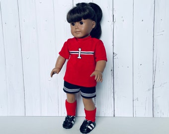 Puede incluir: Una camiseta de fútbol roja con rayas blancas y negras y el número "1" en la parte delantera. La camiseta la lleva una muñeca con el pelo castaño y zapatos de fútbol negros.