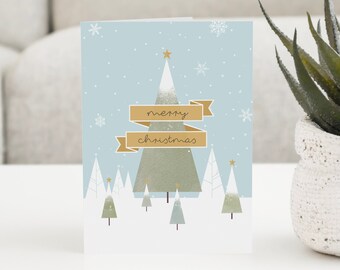 Può includere: Un biglietto di Natale blu con una scena di foresta innevata. Il biglietto presenta un grande albero di Natale verde con una stella dorata in cima e il testo "merry christmas" su uno stendardo dorato.