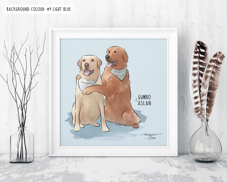 Framed illustration of two dogs, one golden and one yellow, wearing bandanas, with names "Gumbo" and "Aslan" labeled. The frame sits on a white table with minimalistic decor and a light blue background.