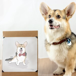 A Corgi sits next to a framed illustration of itself wearing a bow tie. The dog has a collar and a tag, and both it and the illustration are on a wooden surface with a plain background.