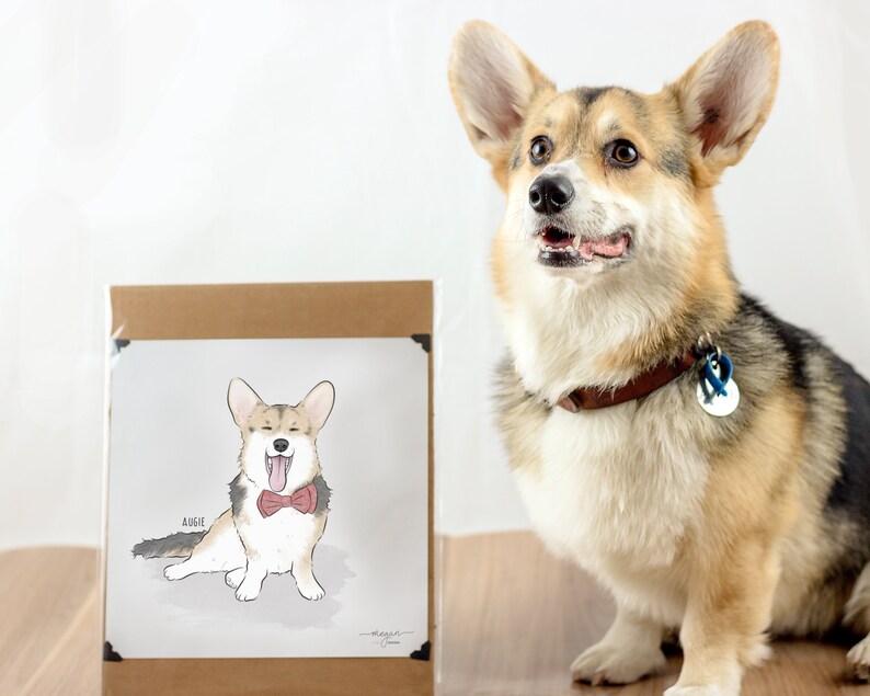A Corgi sits next to a framed illustration of itself wearing a bow tie. The dog has a collar and a tag, and both it and the illustration are on a wooden surface with a plain background.