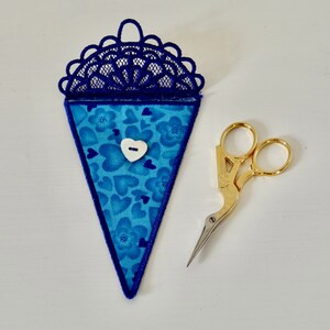 Può includere: Un porta forbici in tessuto blu con un motivo floreale e a cuore, un bottone bianco a forma di cuore e un top in pizzo blu scuro. Le forbici da ricamo color oro sono accanto al porta forbici.