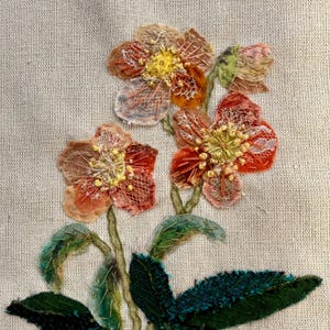 Puede incluir: Primer plano de una obra de arte textil con flores tridimensionales. Las flores son en tonos naranja, melocotón y amarillo con detalles intrincados. Hojas y tallos verdes completan el diseño floral, sobre un fondo neutro.