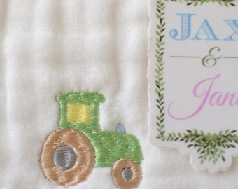 Può includere: Un tessuto bianco con un trattore verde ricamato su di esso. Un'etichetta bianca con un bordo di foglie verdi dice "Jax & Jane" in lettere blu e rosa.