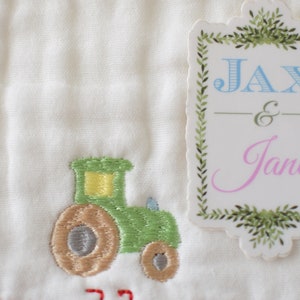 May include: A white fabric with a green tractor embroidered on it. A white tag with a green leafy border says "Jax & Jane" in blue and pink lettering.