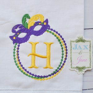 May include: A white embroidered kitchen towel with a purple, green, and yellow Mardi Gras mask and beads surrounding a gold letter H.