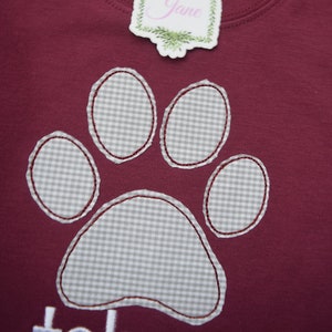 May include: Burgundy red t-shirt with a gray and white gingham paw print design. The shirt has a tag that says "Jax & Jane".