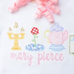 May include: White t-shirt with embroidered images of a candelabra, a rose in a glass dome, a teapot, and a green frame with the text "Jax & Jane". The text "mary pierce" is embroidered in pink below the images.