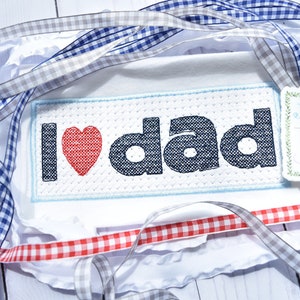 May include: White fabric with a blue and white checkered ribbon, a gray ribbon, and a red and white checkered ribbon. The fabric has an embroidered design that says "I love dad" with a red heart. There is a small tag with the text "Jax & Jane" on it.