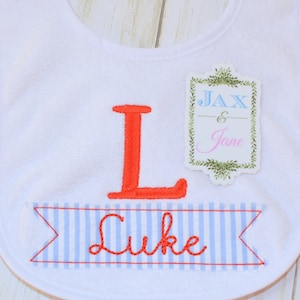 May include: White baby bib with blue and red striped banner that says "Luke" in red embroidery. A separate tag with green leaves and a white background says "Jax & Jane" in blue and pink embroidery.