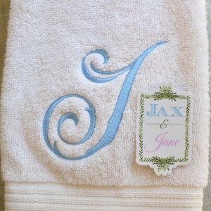 Op de afbeelding: Witte badhanddoek met een blauwe geborduurde letter J en een roze en groen label met de tekst "Jax & Jane".