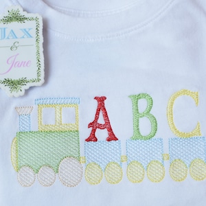 Puede incluir: Camiseta blanca con un diseño de tren bordado y las letras "ABC" en rojo, verde y amarillo. El tren es de colores pastel. Una etiqueta con el texto "Jax & Jane" está adjunta. Se ve una tela a cuadros azul y blanco.