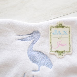 May include: White towel with a blue embroidered pelican and a tag that says "Jax & Jane" in blue and pink lettering.