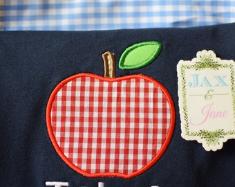 Puede incluir: Una camisa azul marino con una manzana de cuadros rojos y blancos y una etiqueta blanca con el texto "Jax & Jane" en azul y rosa.