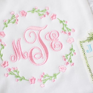 May include: White embroidered baby blanket with a pink floral wreath and the monogram "J.E." in the center. A tag with the text "Jax & Jane" is attached to the blanket.