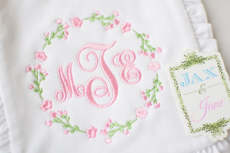 May include: White embroidered baby blanket with a pink floral wreath and the monogram "J.E." in the center. A tag with the text "Jax & Jane" is attached to the blanket.
