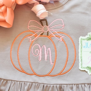 Pode incluir: Uma blusa cinza claro com um desenho de abóbora laranja bordada, um laço rosa e a letra "M". A blusa tem uma bainha com babados e uma etiqueta que diz "Jax & Jane". Um laço cor de pêssego está no canto superior esquerdo.