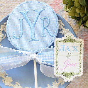 May include: A blue and white gingham circle with the monogram "JYR" embroidered on it, attached to a white stick. A blue and white ribbon is tied around the stick. A white card with green foliage border and the text "Jax & Jane" in pink and blue lettering.