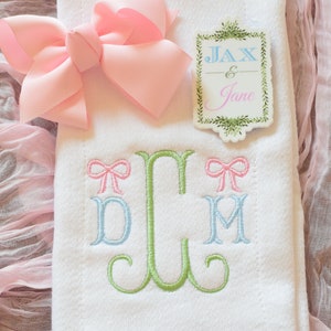 May include: White embroidered towel with a green monogram and two pink bows. The monogram is "JCM". A pink bow is on each side of the monogram. The towel is on a pink fabric background. A sticker with the text "Jax & Jane" is on the towel.