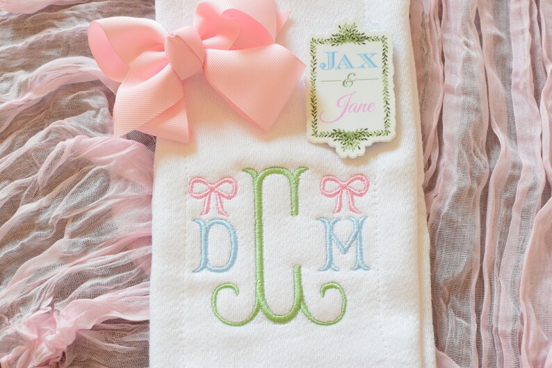 May include: White embroidered towel with a green monogram and two pink bows. The monogram is "JCM". A pink bow is on each side of the monogram. The towel is on a pink fabric background. A sticker with the text "Jax & Jane" is on the towel.