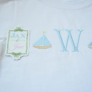 May include: White t-shirt with blue and yellow embroidered sailboats and the letter "W" in blue. A tag with the text "Jax & Jane" is attached to the shirt.