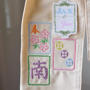 May include: Cream-colored fabric with embroidered designs. Features floral and Chinese character motifs in pink, orange, and purple, framed in colored borders. Includes a sticker with the names "Jax & Jane" in blue and pink.