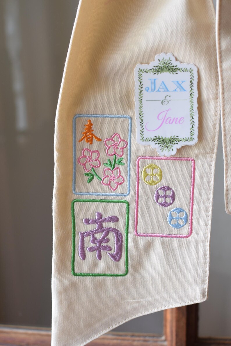 May include: Cream-colored fabric with embroidered designs. Features floral and Chinese character motifs in pink, orange, and purple, framed in colored borders. Includes a sticker with the names "Jax & Jane" in blue and pink.