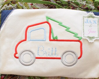 May include: Cream-colored Christmas-themed pillow with an embroidered red truck carrying a green Christmas tree. The truck has light blue accents and the name "Bill" embroidered in light blue. A tag with the text "Jax & Jane" is attached.