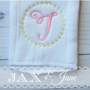 May include: A white cloth burp cloth with a pink embroidered letter T inside a yellow dotted circle. The text "JAX - & - Jane" is at the bottom of the image.