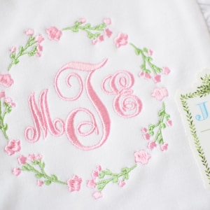 May include: White embroidered baby blanket with a pink floral wreath and the monogram "J.S." in the center. A tag with the text "Jax & Jane" is attached to the blanket.