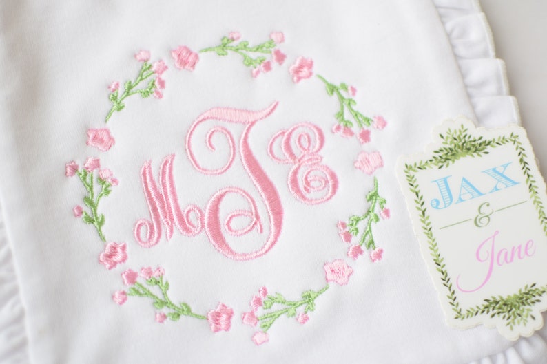 May include: White embroidered baby blanket with a pink floral wreath and the monogram "J.S." in the center. A tag with the text "Jax & Jane" is attached to the blanket.