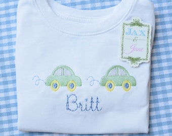 Puede incluir: Un body blanco para bebé con coches bordados en verde y el nombre "Britt" cosido debajo. Una etiqueta con el texto "Jax & Jane" está unida al body.