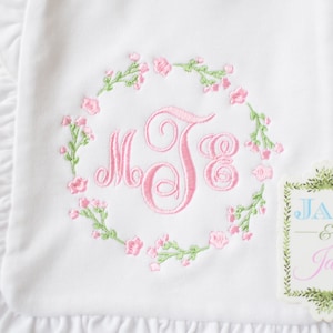 May include: White embroidered baby burp cloth with pink floral wreath and monogram "MJB". The cloth has a ruffled edge and a small tag that reads "Jax & Jane".