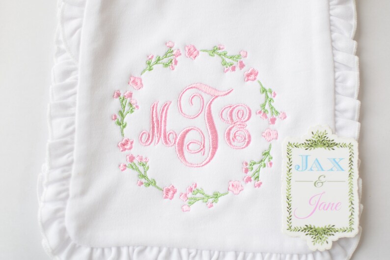 May include: White embroidered baby burp cloth with pink floral wreath and monogram "MJB". The cloth has a ruffled edge and a small tag that reads "Jax & Jane".