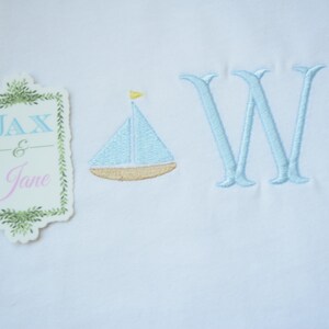May include: A white fabric with blue and yellow embroidered sailboats and the letter "W". A tag with the text "Jax & Jane" is in the upper left corner.