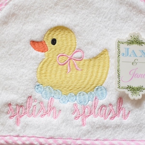 May include: White hooded baby towel with a yellow embroidered duck wearing a pink bow, surrounded by blue bubbles. The words "splish splash" are embroidered in pink. A tag with the names "Jax & Jane" is on the right.