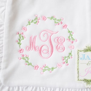 May include: White baby blanket with a pink embroidered monogram surrounded by a wreath of pink flowers and green leaves. The blanket has a ruffled edge. A tag with the text "JAX & Jane" is attached to the blanket.