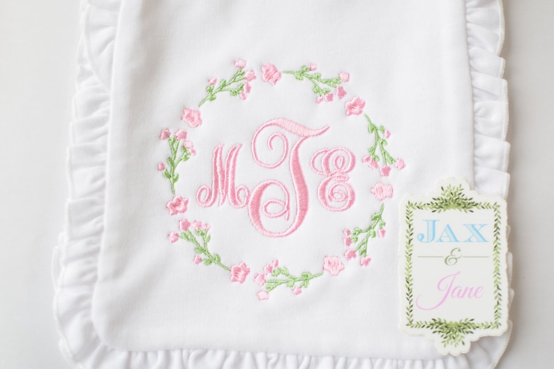 May include: White baby blanket with a pink embroidered monogram surrounded by a wreath of pink flowers and green leaves. The blanket has a ruffled edge. A tag with the text "JAX & Jane" is attached to the blanket.