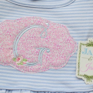 May include: A light blue and white striped baby outfit with a floral patch featuring the letter "G". The patch is pink with a floral pattern and a light blue embroidered letter. A tag with the text "Jax & Jane" is attached.