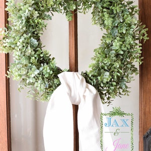 May include: A green leafy wreath with a white fabric bow tied around it. The wreath is hanging on a wooden door with a glass pane. The text "JAX & Jane" is written on a sign in the bottom right corner of the image.