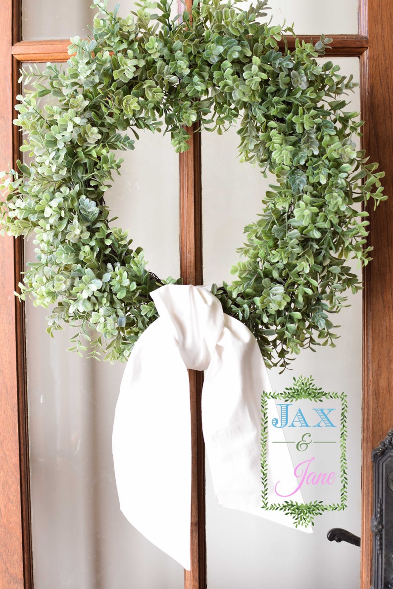 May include: A green leafy wreath with a white fabric bow tied around it. The wreath is hanging on a wooden door with a glass pane. The text "JAX & Jane" is written on a sign in the bottom right corner of the image.