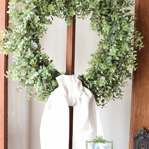 Peut inclure: Une couronne de feuilles vertes avec un nœud en tissu blanc suspendu à une porte en bois avec un panneau en verre. La couronne porte le texte "Jax & Jane" dans un cadre de feuilles vertes.