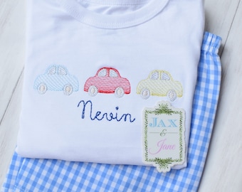 Puede incluir: Un pelele blanco con coches bordados en azul, rojo y amarillo. El pelele tiene el nombre "Nevin" bordado. Un tejido vichy azul y blanco es visible debajo del pelele. Una etiqueta blanca con hojas verdes alrededor del borde tiene el texto "JAX & Jane".