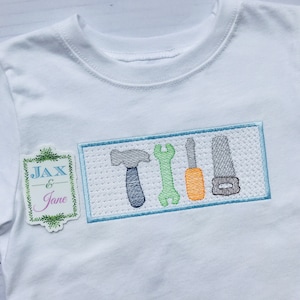Puede incluir: Camiseta blanca con un diseño bordado en azul, verde y naranja de herramientas en un rectángulo. El texto "Jax & Jane" está bordado en una etiqueta verde y blanca.