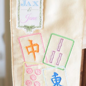 May include: Cream-colored fabric with embroidered Mahjong tiles and a decorative label that reads "Jax & Jane". The Mahjong tiles are in various colors, including orange, green, pink, and blue.