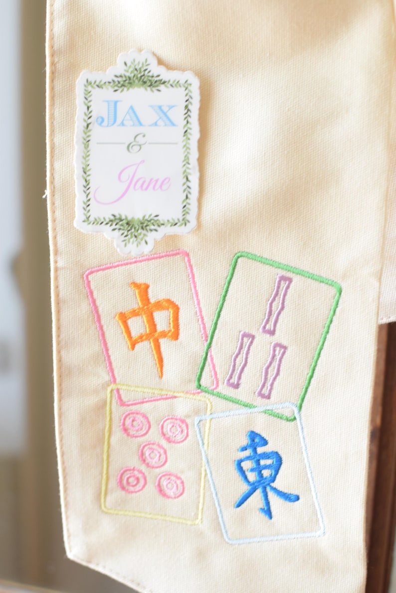 May include: Cream-colored fabric with embroidered Mahjong tiles and a decorative label that reads "Jax & Jane". The Mahjong tiles are in various colors, including orange, green, pink, and blue.